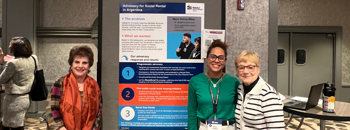 El poster de Hábitat para la Humanidad Argentina y tres mujeres paradas frente al mismo sonriendo.