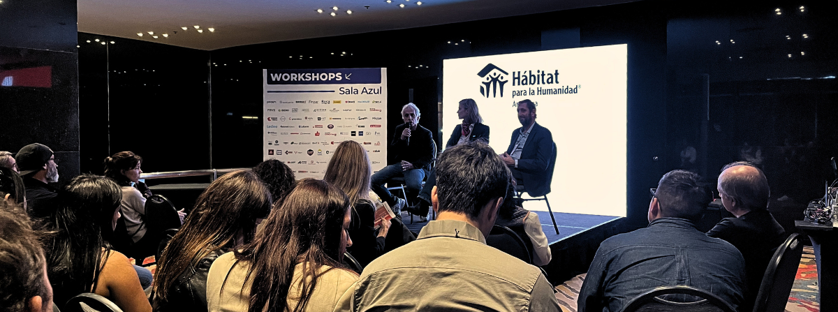 Panel de tres ponentes sentados en un escenario durante un workshop en la Sala Azul, con una pantalla proyectando el logo de Hábitat para la Humanidad Argentina, frente a una audiencia sentada en una sala de conferencias con iluminación tenue